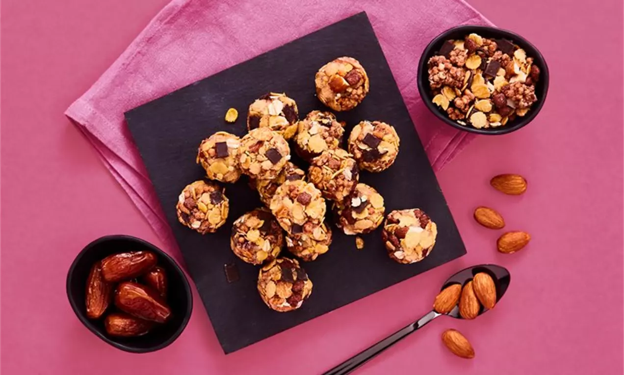 Okrogle proteinske kroglice v zlatorjavi teksturi s sadnimi koščki in oreščki na črni skrilasti plošči s skledami datljev in granole