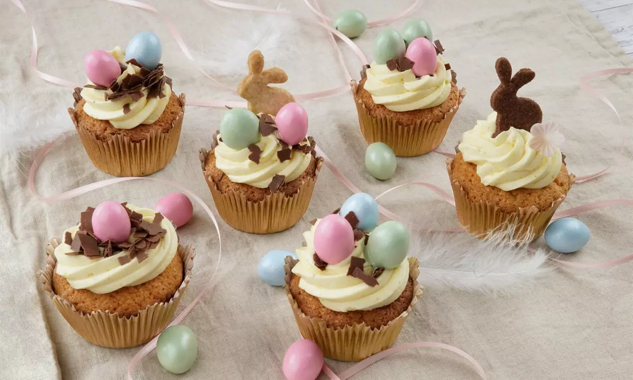 Ванилови къпкейкове със светъл маслен крем, украсени с цветни мини яйца, шоколадови стърготини и захарни фигурки върху ленена покривка