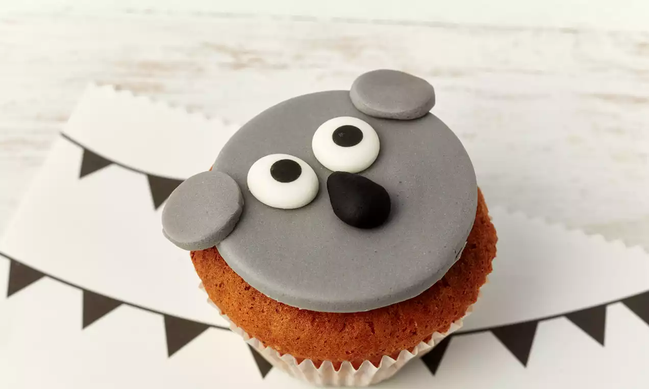Muffin mit goldbraunem Teig und grauer Fondantdecke in Form eines Koalagesichts mit schwarzen und weißen Details auf Papier mit Wimpelmuster