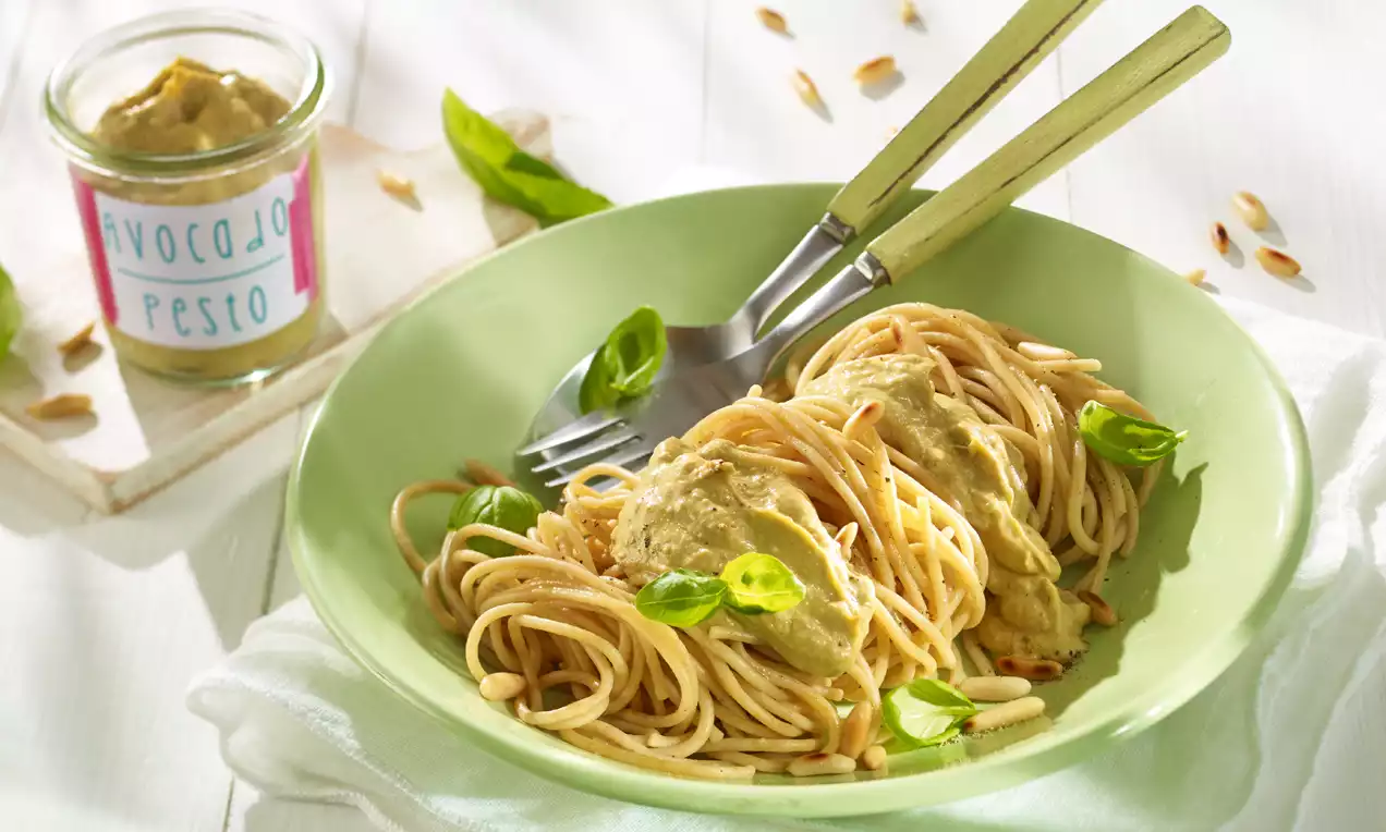 Spaghetti mit cremigem Avocado-Pesto, angerichtet auf einem grünen Teller, garniert mit frischen Basilikumblättern und gerösteten Pinienkernen
