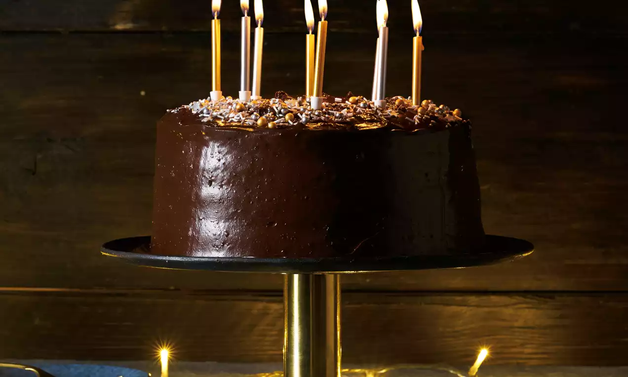 Chocolate birthday cake with glossy buttercream, topped with chocolate shavings and lit gold and silver tall candles on a dark pedestal stand