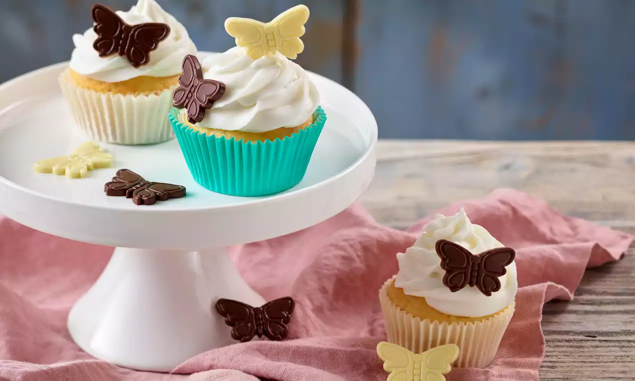 Vanille-Muffins mit weißem Sahnetopping, verziert mit Schoko-Schmetterlingen in Dunkelbraun und Weiß, auf weißer Tortenplatte und rosa Stoff