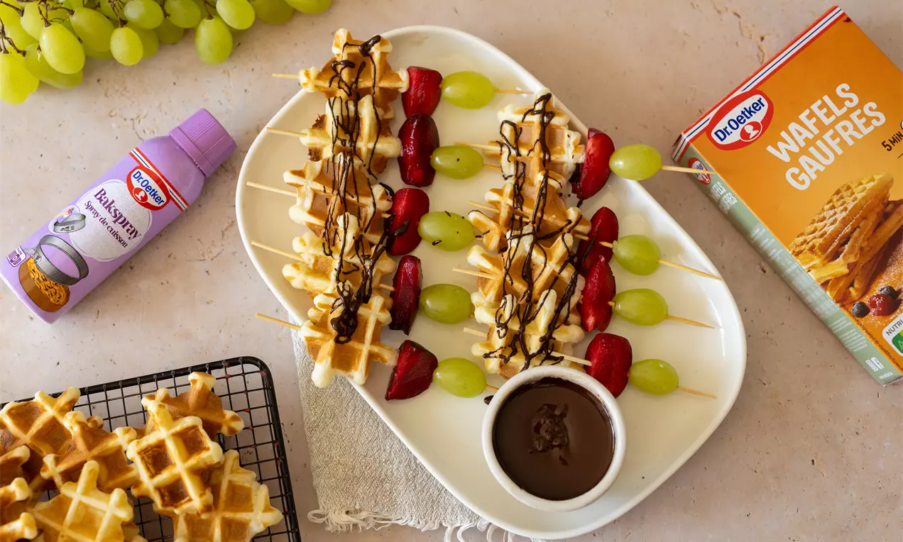 Wafelbrochettes met stukjes wafel, rode aardbeien en groene druiven op houten prikkers, gegarneerd met chocoladesaus op een witte schaal