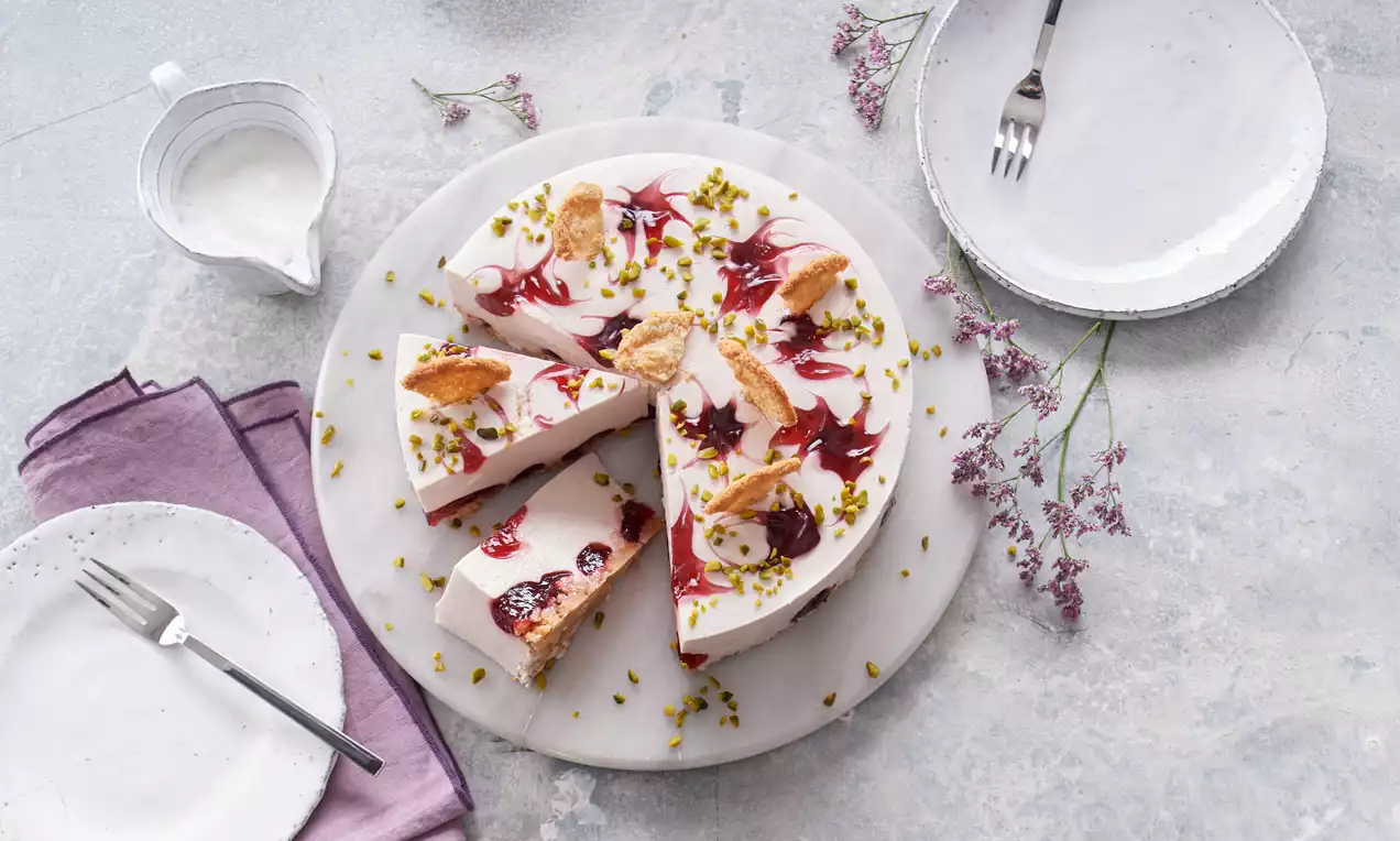 Buttermilch-Frischkäsetorte mit Cassis auf weißem Teller, verziert mit roten Cassis-Swirls, Pistazien und Keksstücken, umgeben von lila Blüten