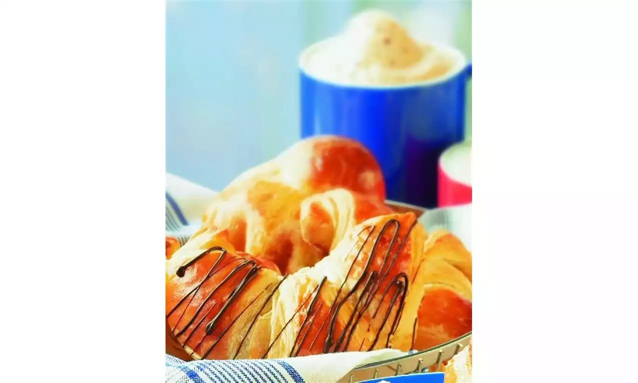 Croissants dorés et moelleux nappés de fines filets de chocolat noir, disposés dans un panier garni d'un tissu rayé bleu et blanc avec une tasse mousseuse en arrière-plan