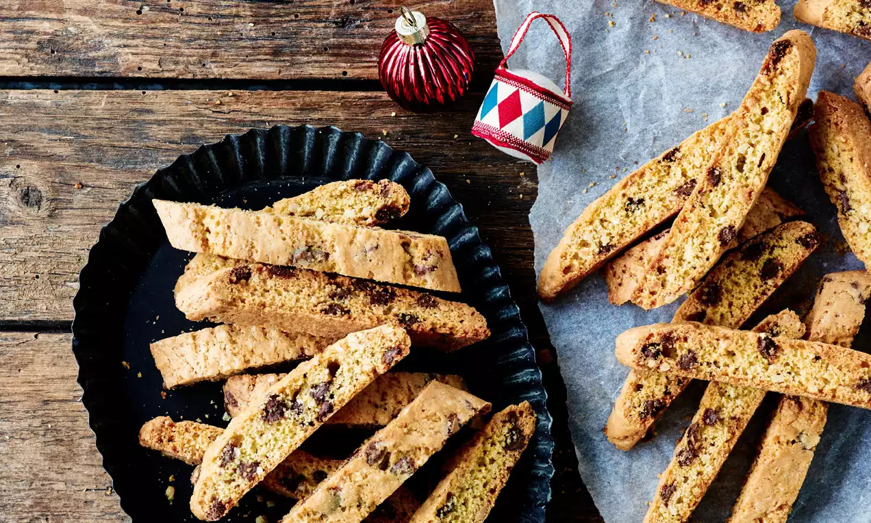 Lysebrune biscotti med sprø overflate og små sjokoladebiter, noen plassert på sort, riflet fat, andre på bakepapir ved siden av