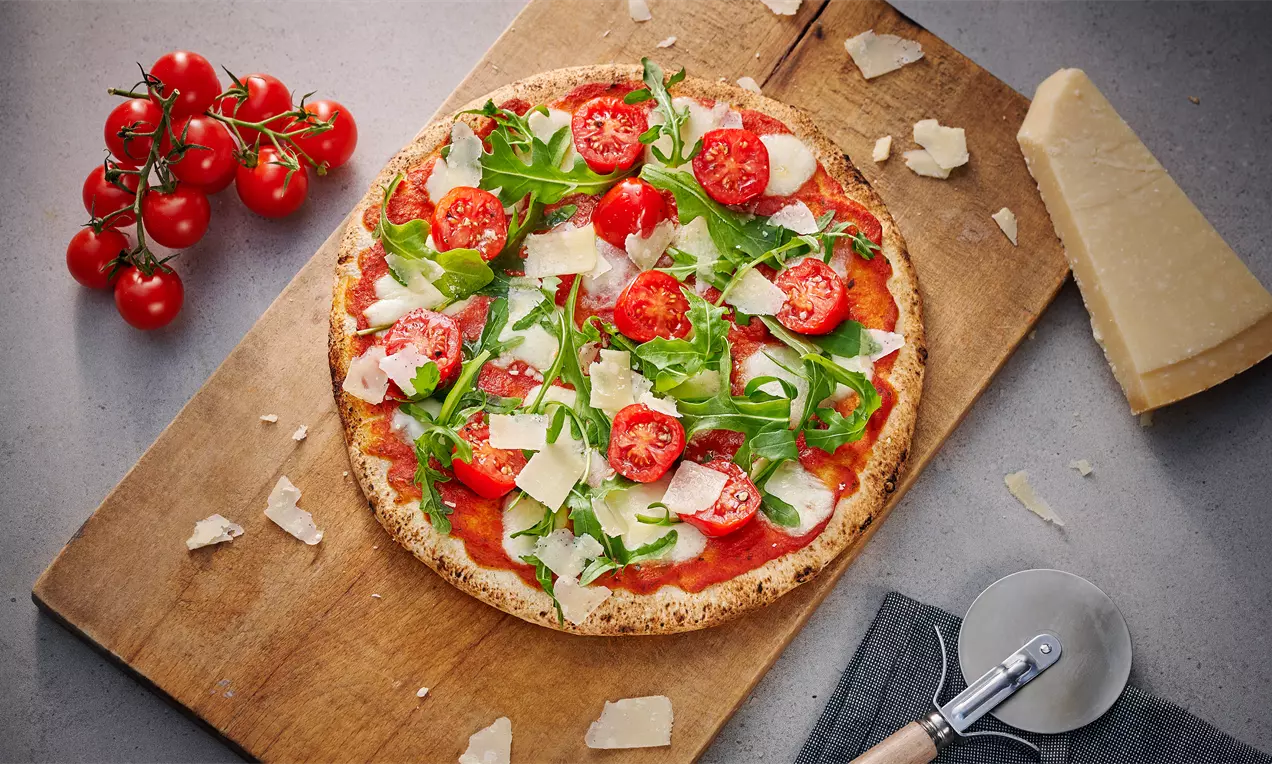 Pizza de masa fina sobre tabla de madera, cubierta con salsa roja brillante, rodajas de tomate, rúcula fresca y lascas de queso parmesano