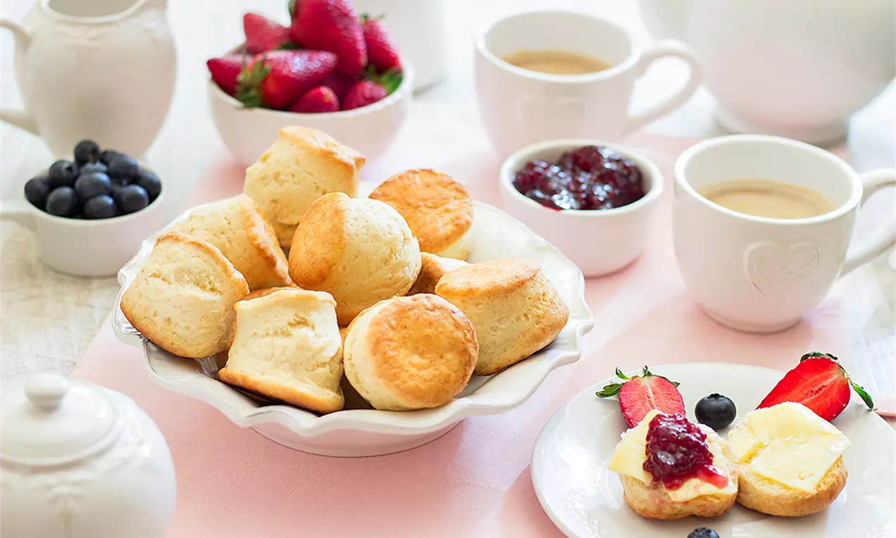 Златисто-кафяви домашни бисквити с кръгла форма и мека текстура, подредени в бяла купа на маса с плодове, чай и конфитюр