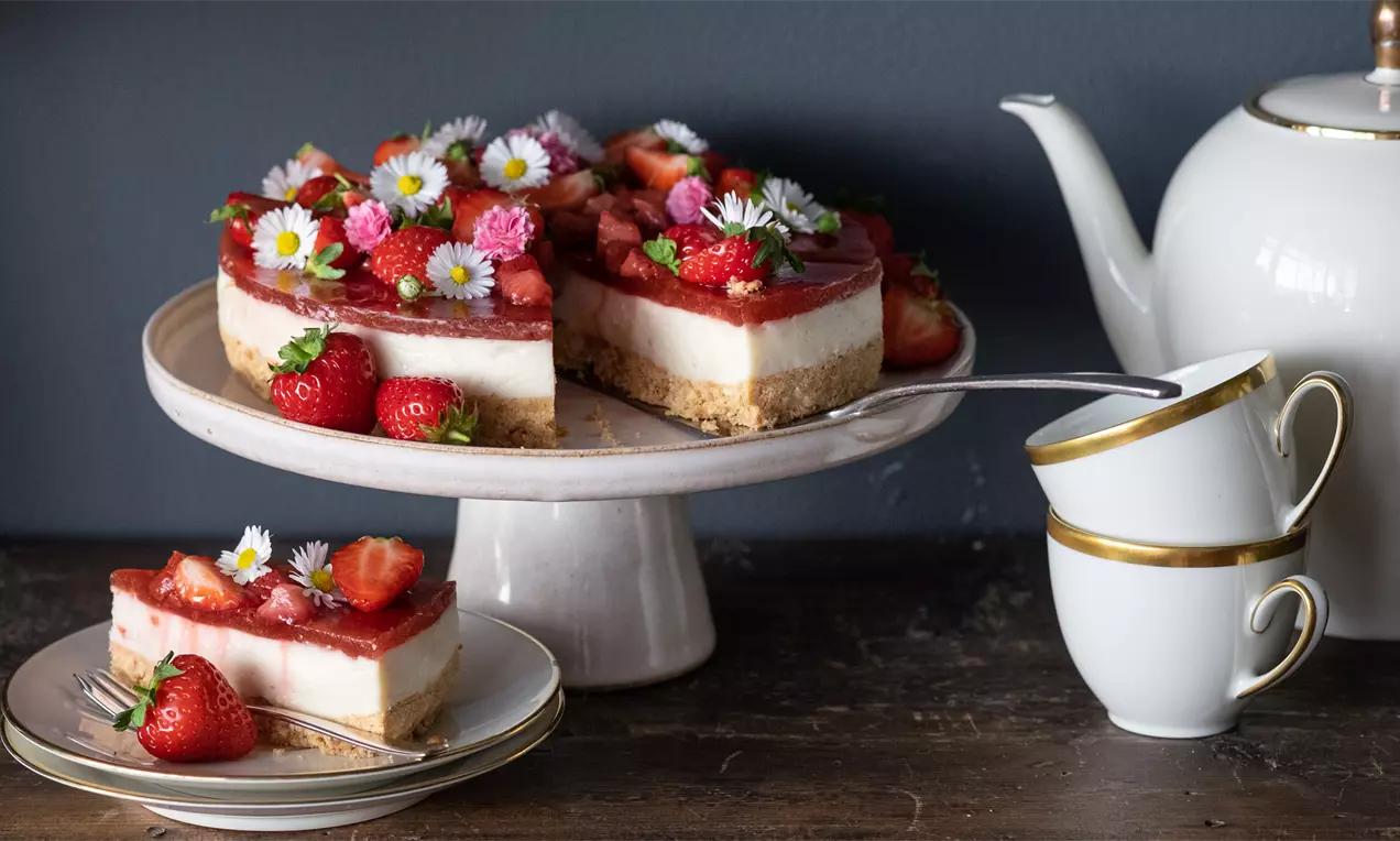 Ягодов чийзкейк с кремообразен бял слой върху хрупкав бисквитен блат, покрит с червен ягодов топинг, свежи ягоди и цветчета върху порцеланова поставка