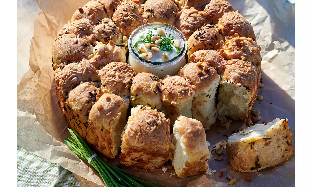 Kerekre formált, pirosas-aranyhéjú, rusztikus kenyérdarabokból álló gyűrű, közepén krémes mártogató kicsi tálban, apró zöld díszítéssel