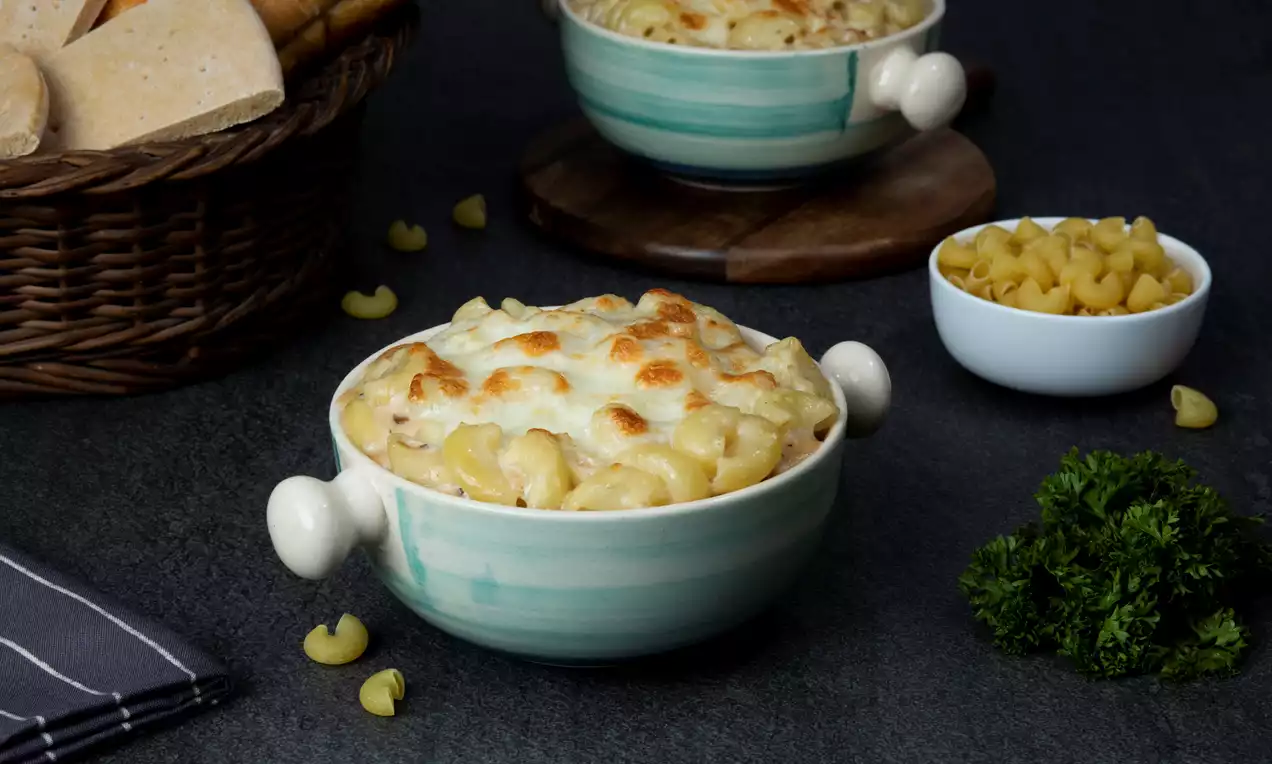 Creamy macaroni pasta baked in a pale green bowl with golden melted cheese topping, surrounded by parsley, bread, and loose pasta