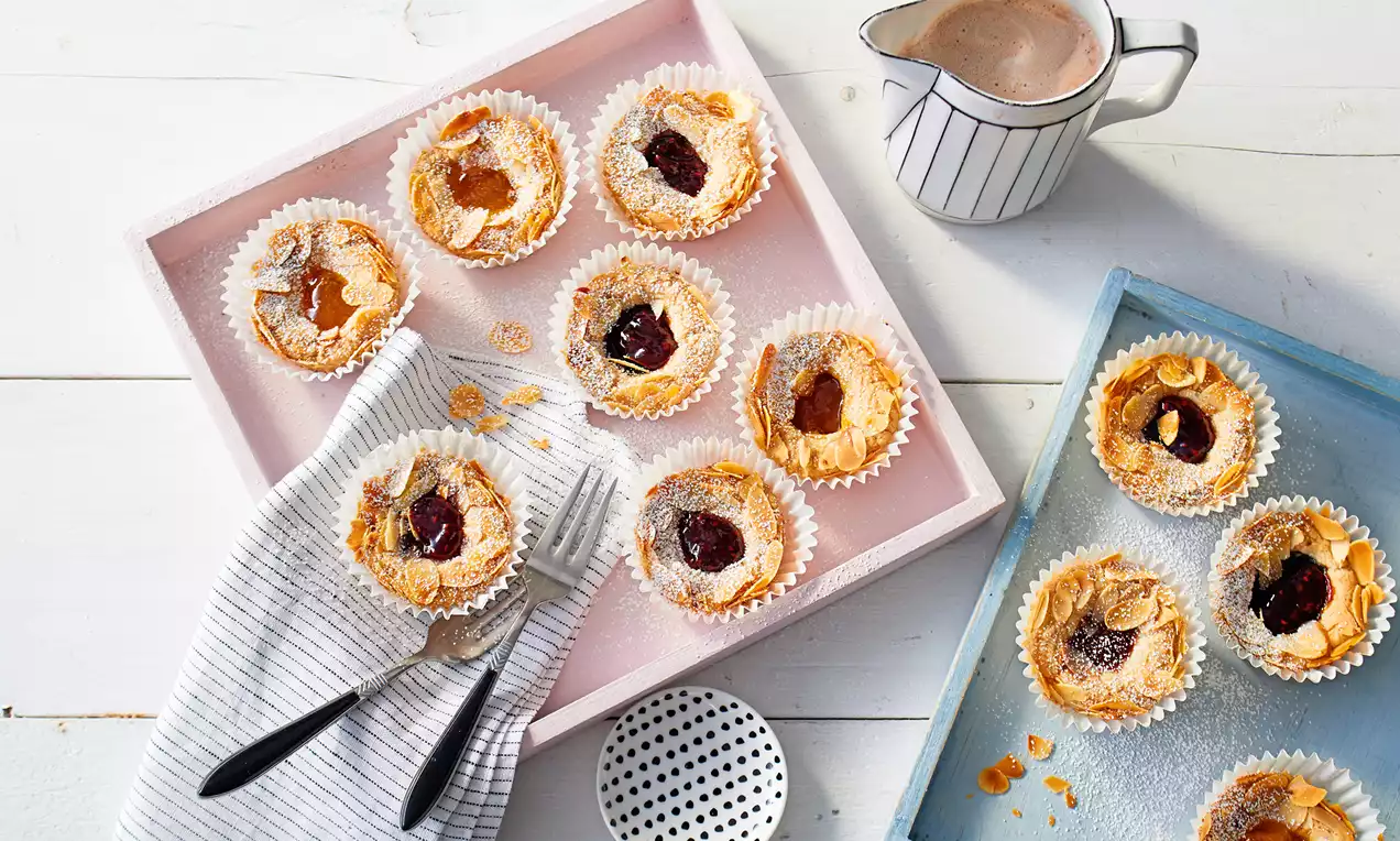 Goldene, filigran gebackene Marmeladen-Küchlein mit leichter Zuckerschicht, garniert mit gerösteten Mandeln in Papierförmchen auf einem blauen Tablett