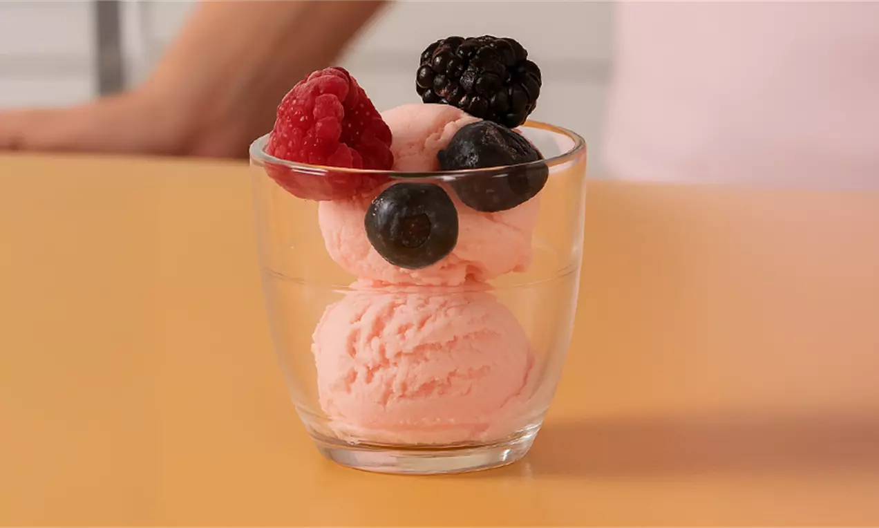 Helado de fresa rosa claro en bolas suaves sobre un vaso transparente, coronado con frambuesas, moras y arándanos vibrantes