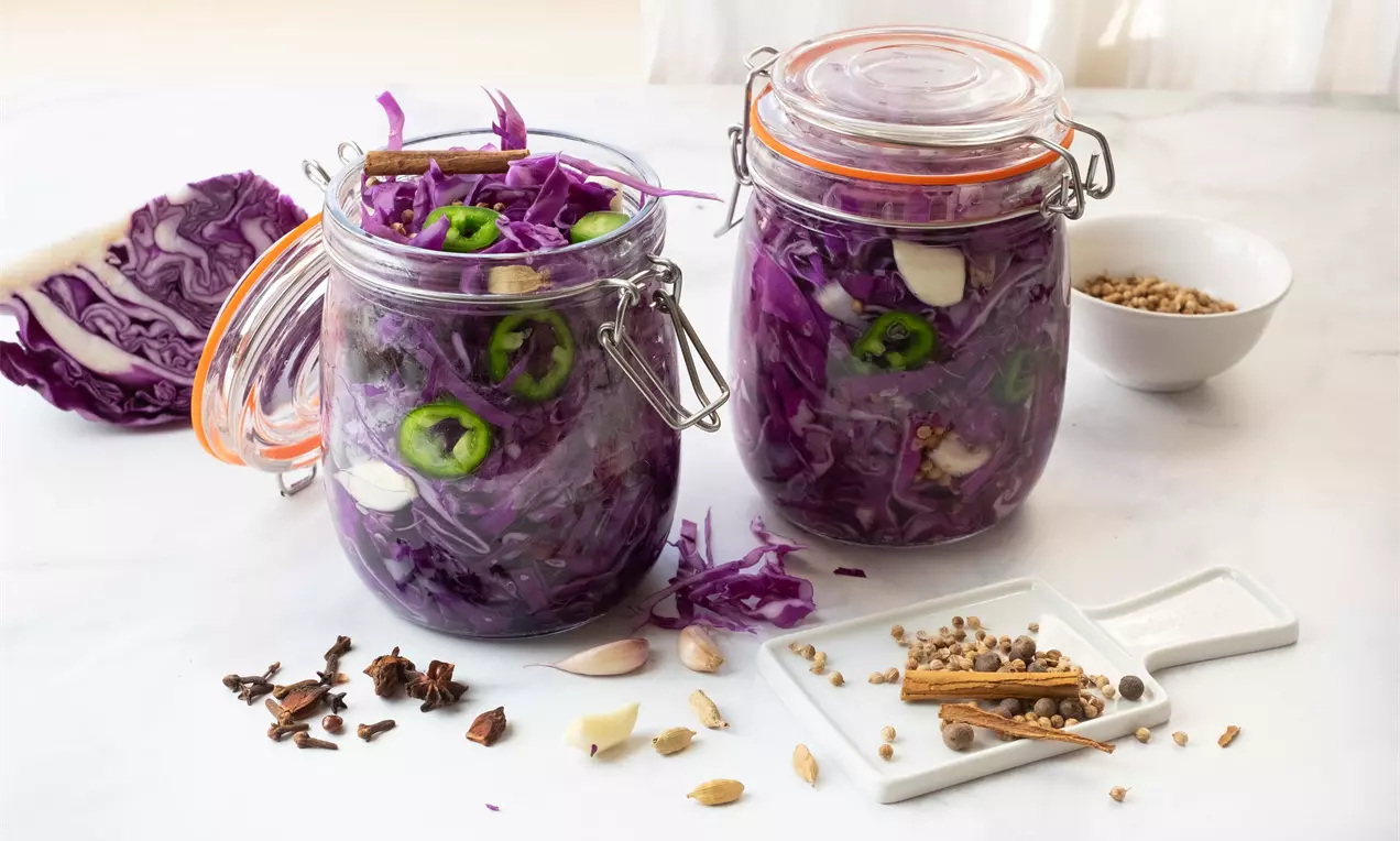 Мариновано ярко лилаво червено зеле на тънки резени със зелени люти чушки и бели скилидки чесън в стъклени буркани на светла повърхност