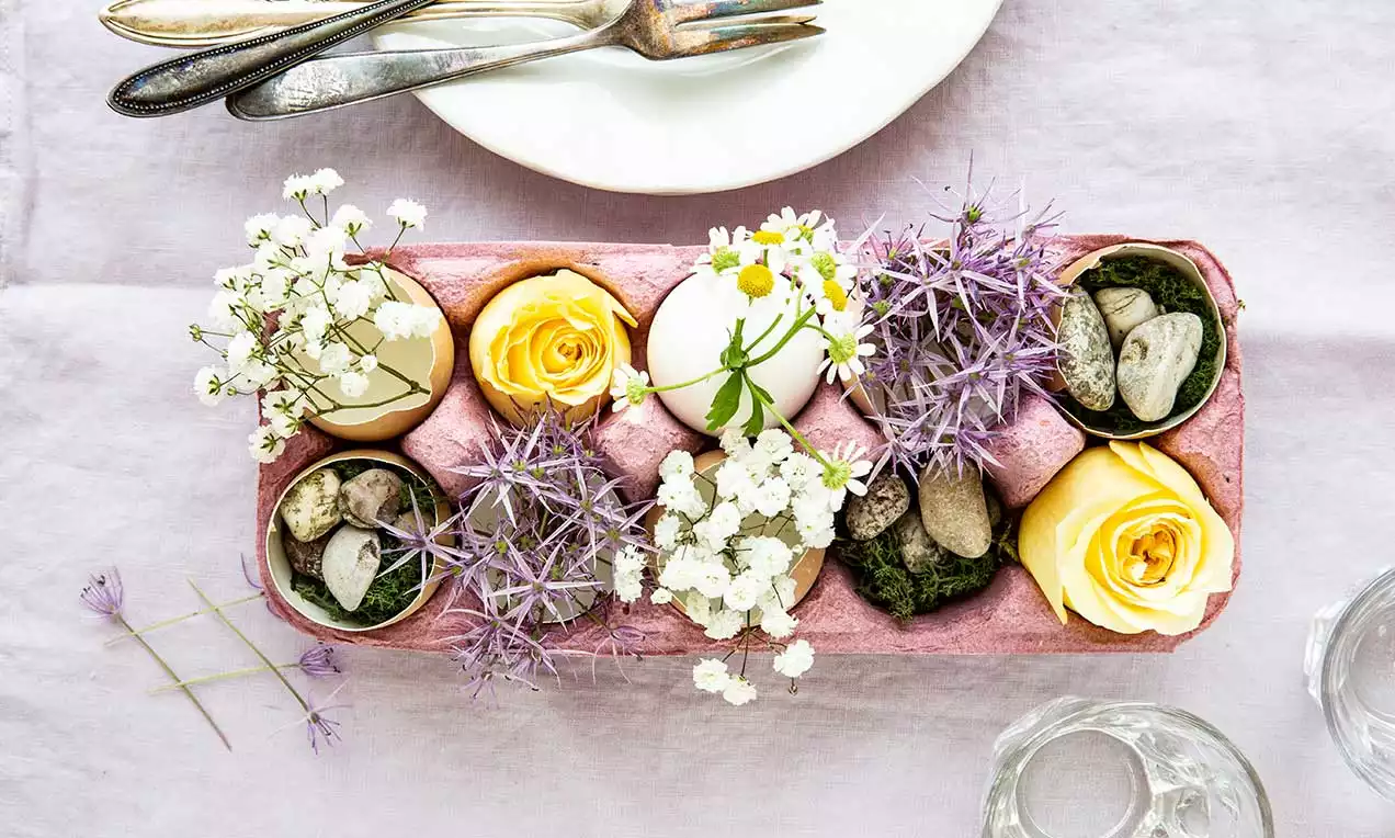 Blassrosa Eierkarton mit gelben Rosen, weißen Blüten, lila Disteln, Moos und Steinen dekoriert auf lila Stofftischdecke drapiert