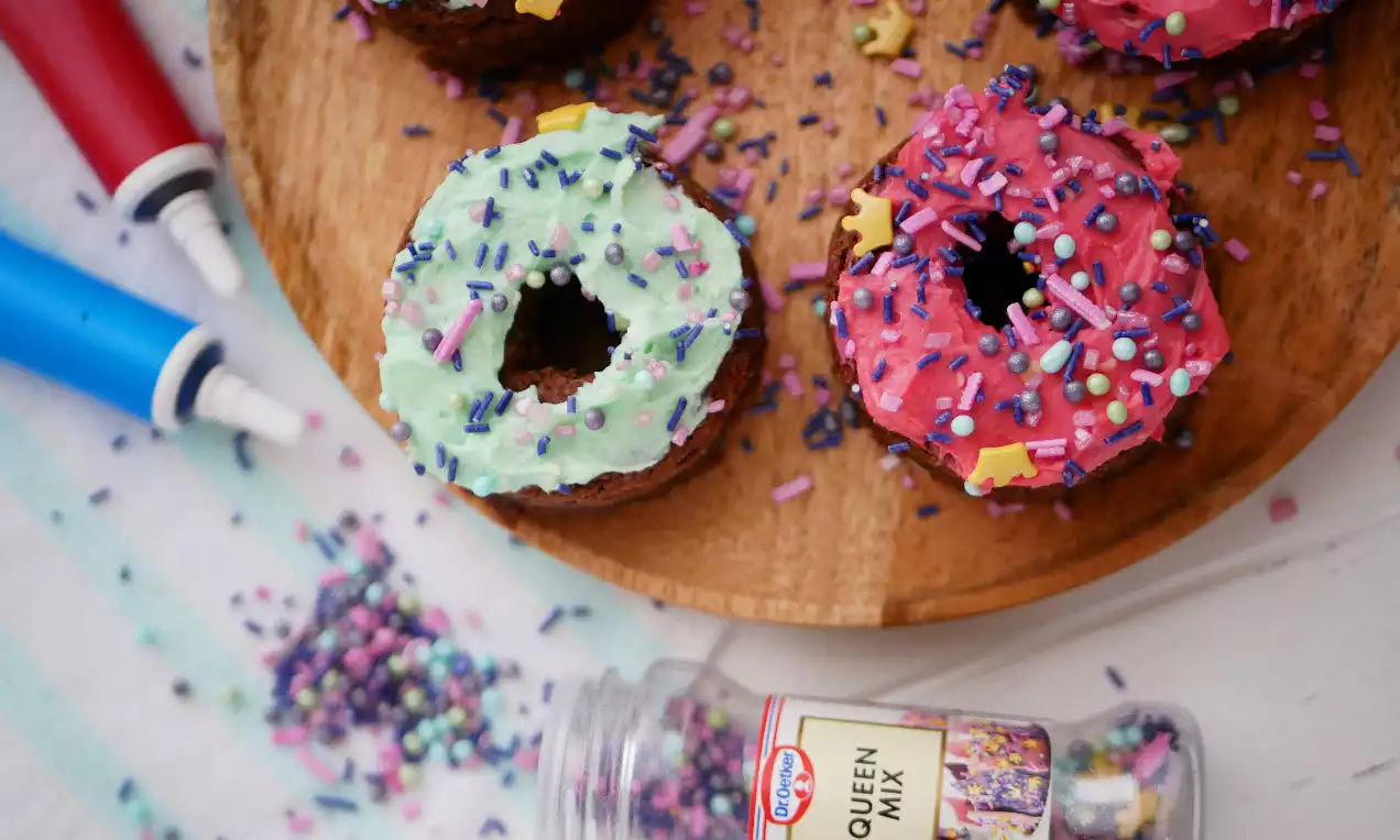 Runda chokladdonuts med grön och rosa glasyr, dekorerade med färgglatt strössel, upplagda på ett träfat med strössel runtom