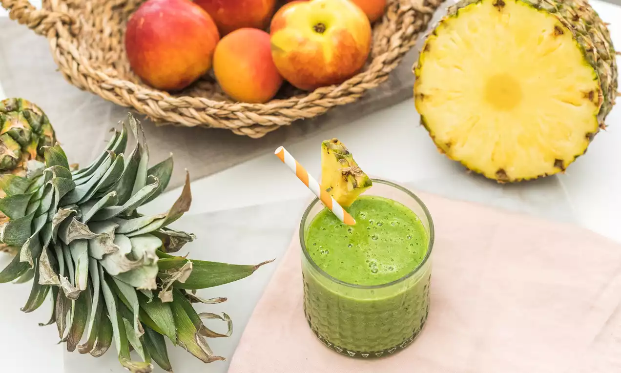 Batido verde brillante en vaso transparente con rodaja de piña y pajitas de rayas, sobre servilleta rosa junto a frutas frescas y media piña
