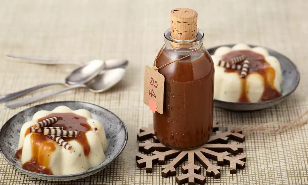 Braune Toffeesoße in einer kleinen Glasflasche mit Korkverschluss, arrangiert auf einem Holz-Schneeflocken-Untersetzer neben zwei Puddingdesserts