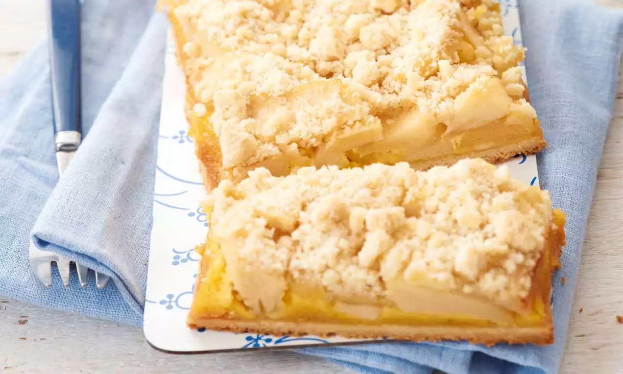 Rechteckiger Apfel-Streuselkuchen mit goldbraunem Boden, saftigen Apfelstücken und feinen helle Streuseln auf einer weißen Platte mit blauem Muster