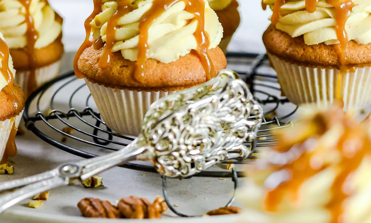 Picture - Caramel and pecan cupcakes with fluffy cream swirls, drizzled caramel sauce and crushed pecans on a cooling rack with scattered nuts