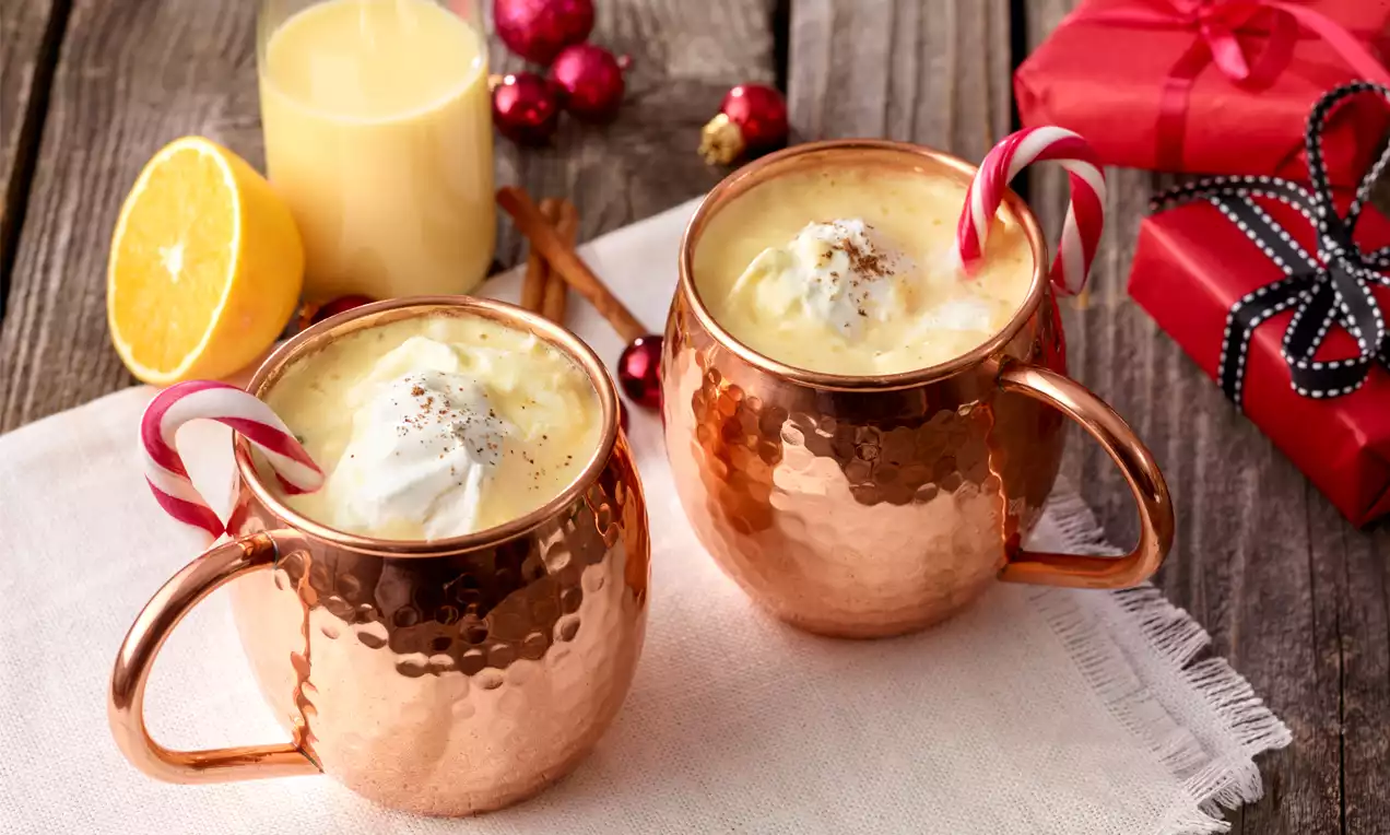 Cremiger Weißwein-Orangen-Punsch in glänzenden Kupferbechern, garniert mit Sahnehaube, Zimt und rot-weißem Zuckerstab auf heller Serviette