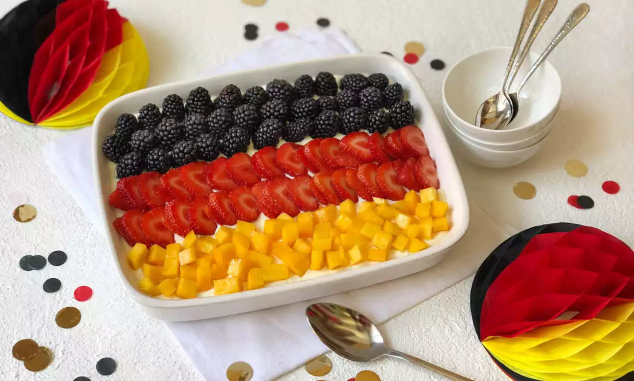 Schichtdessert in rechteckiger weißer Form, belegt mit schwarzen Brombeeren, roten Erdbeerscheiben und gelben Mangowürfeln