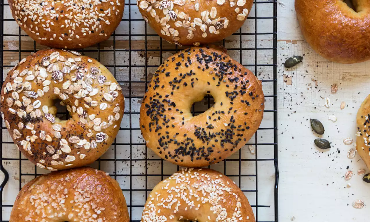 Bagels dorés et brillants, aux graines de sésame, pavot et avoine, disposés sur une grille noire avec des graines éparpillées