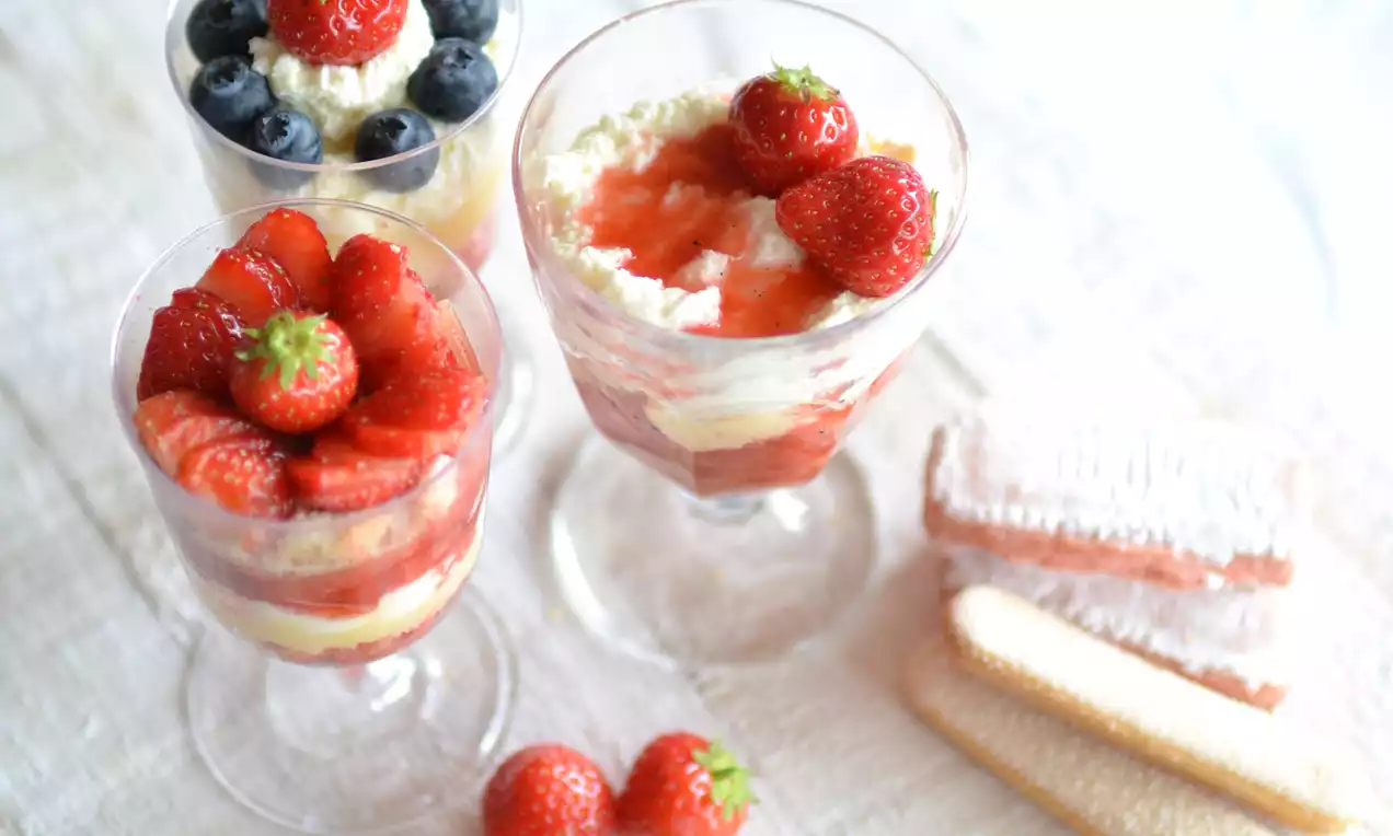 Coupes en verre remplies de crème blanche, génoise légère, morceaux de fraises rouges brillantes et myrtilles sur nappe beige avec biscuits saupoudrés