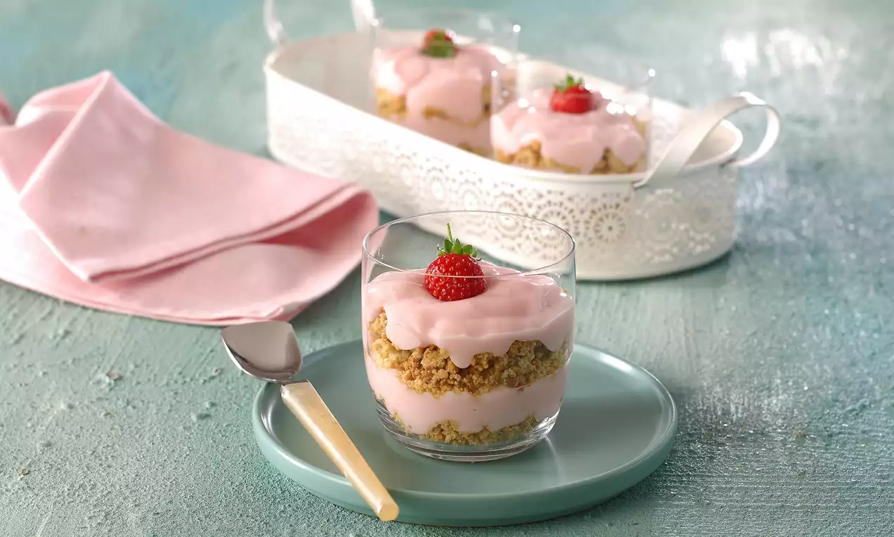 Cam kup içinde kıtır kaju tabanı üzerine pembe çilekli krema, ortada taze çilek, dekoratif tepsi ve pastel tonlarda bir sunum