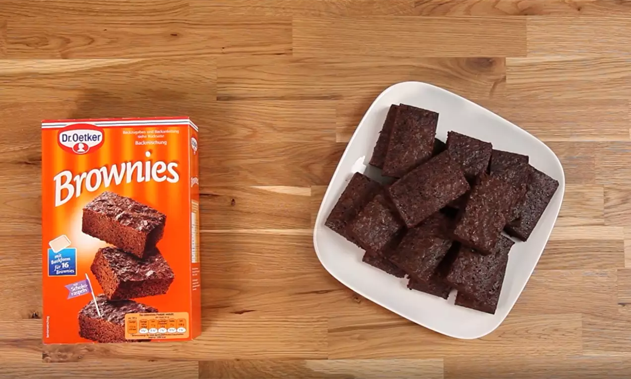 Правоъгълни парчета тъмнокафяво брауни с плътна текстура, наредени куповидно върху бяла квадратна чиния на дървен плот