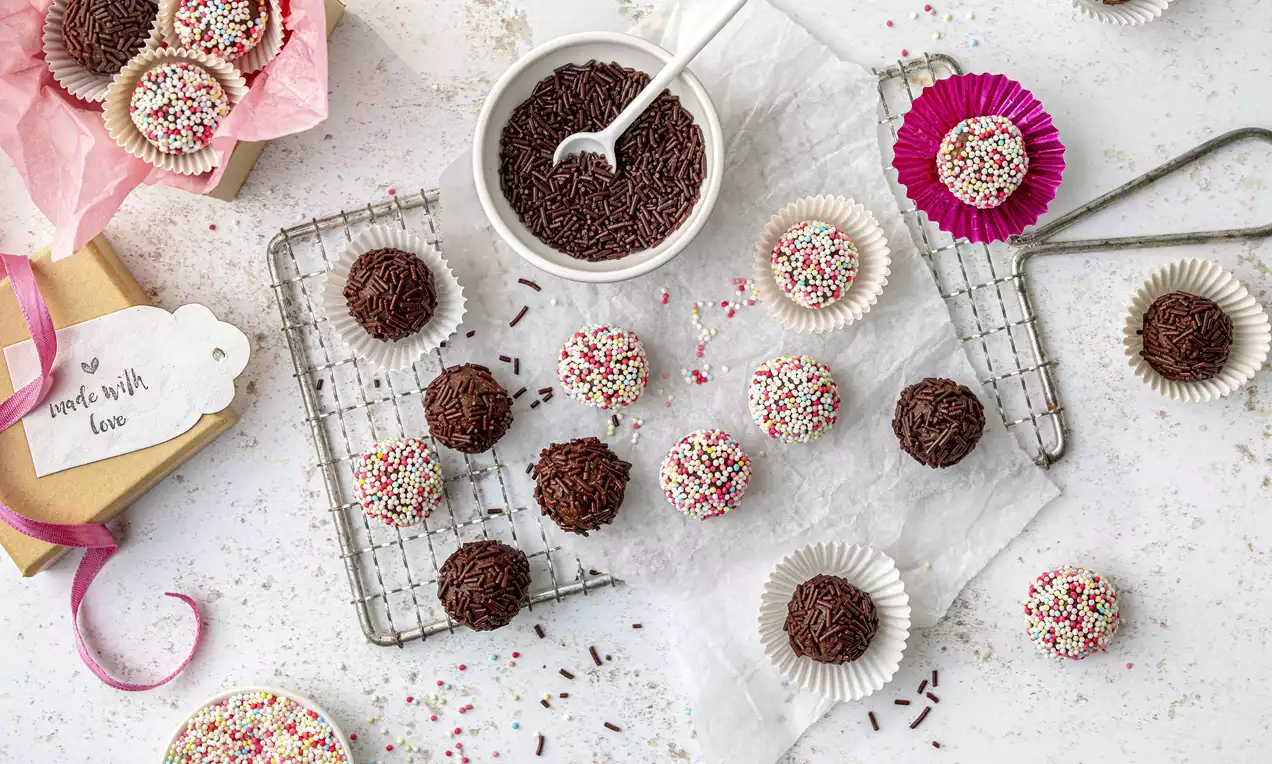 Vegane Pralinen in dunkler Schokolade und bunter Streuseloptik, teils in Papierförmchen und auf einem Gitter mit Dekoration arrangiert