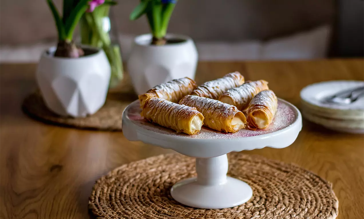 Хрупкави фунийки от златисто тесто с пудра захар, подредени върху бяла керамична поставка, заобиколена от естествени текстури и цветя