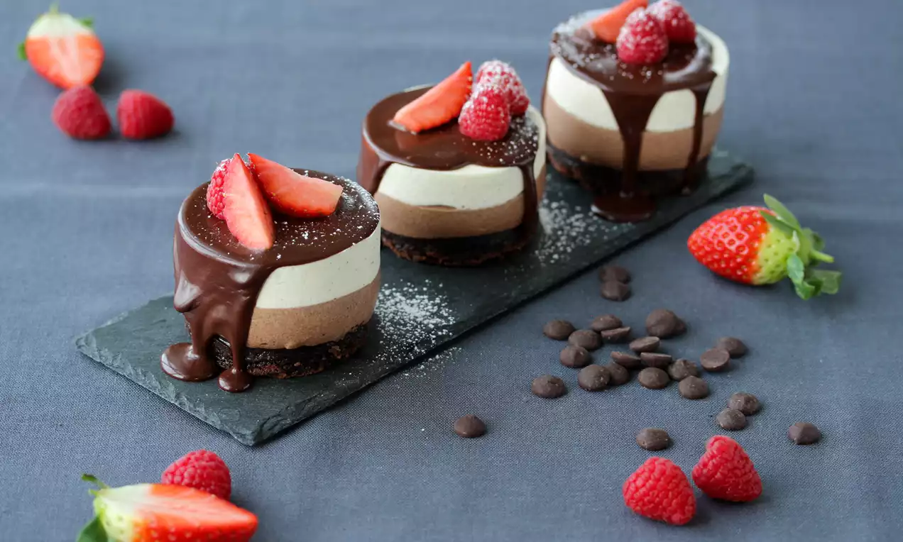 Dessert med lag av brun browniebunn, lys mousse og sjokoladetrekk, toppet med bringebær, jordbærskiver og melisdryss på mørk skiferplate