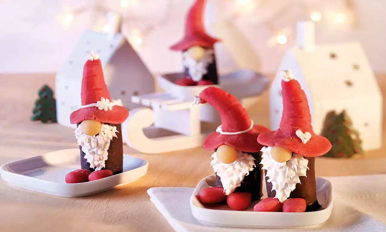 Petits gâteaux chocolatés en forme de lutins avec bonnets rouges en pâte d'amande, barbes en crème blanche et détails décoratifs sur des assiettes blanches