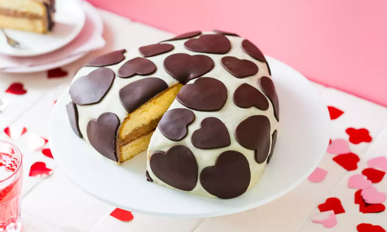 Hartvormige biscuittaart met een witte glazuurlaag, versierd met bruine chocolade harten, op een wit bord met rode confetti