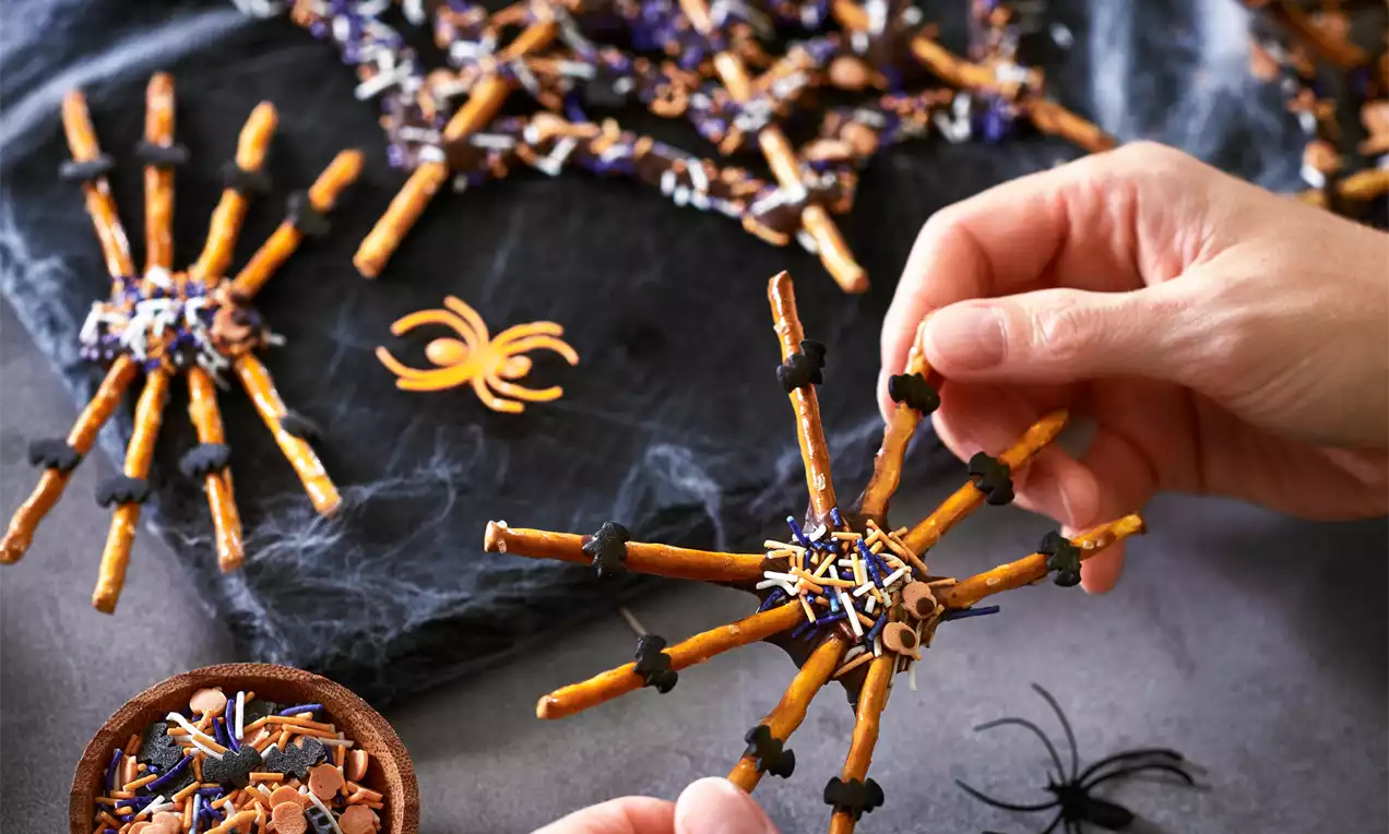 Red de telarañas comestible de pretzels dorados unidos con chocolate negro, decorados con chispas moradas, naranjas y blancas sobre fondo oscuro