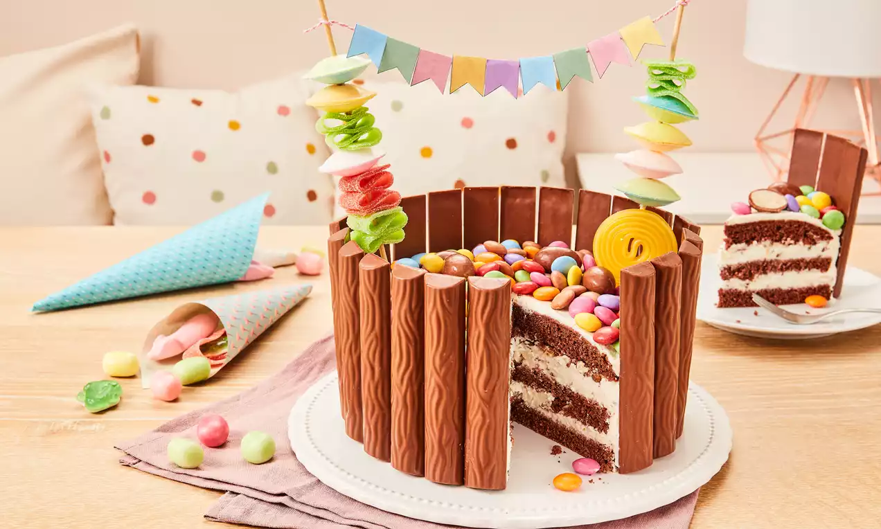 Kleine runde Torte mit Schokoladenstäben umrandet, bunter Süßigkeitenfüllung, Zuckerdekor und Wimpelgirlande auf weißem Tortenteller