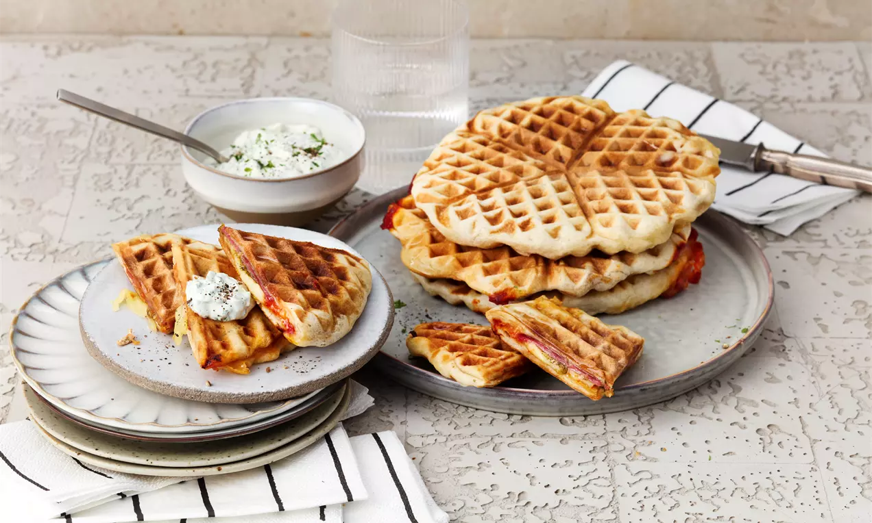 Goldbraune, herzförmige Waffeln mit gefüllter Mitte, gestapelt und angeschnitten, auf Tellern mit Kräuterdip und Servietten drapiert