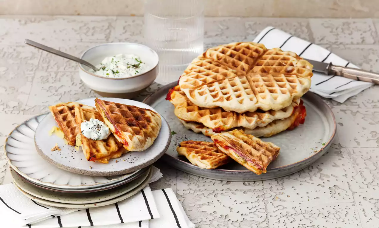 Pizzawaffeln goldbraun mit rautenförmiger Textur, auf einem Teller gestapelt, daneben Stücke mit Kräuter-Dip auf hellem Untergrund