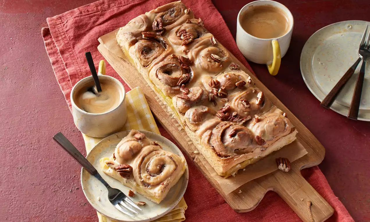 Goldbrauner Zimtschnecken-Apfelkuchen auf einem Holzbrett, garniert mit gehackten Pekannüssen, begleitet von zwei Tassen Kaffee und Teller mit Besteck