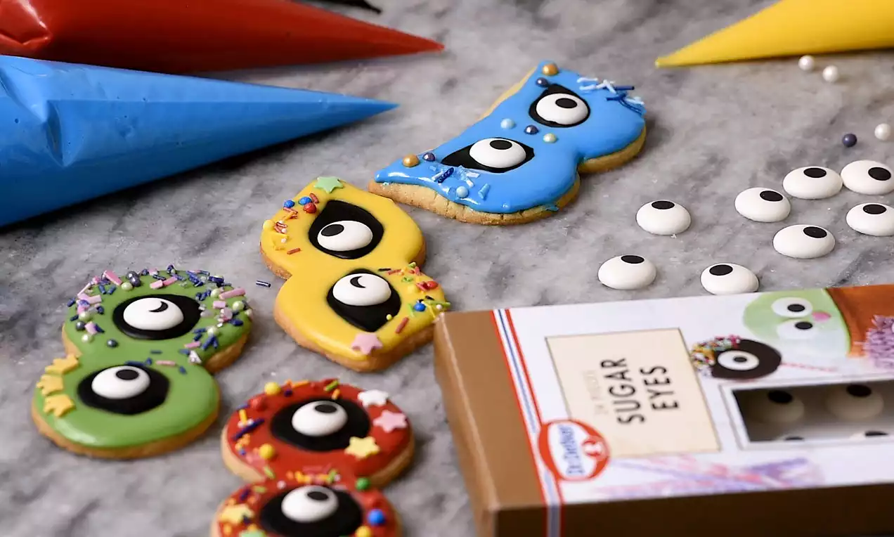 Fastelavnsboller formet som farverige masker i grøn, rød, gul og blå med glasur, sukkerøjne og krymmel på en marmorbordplade