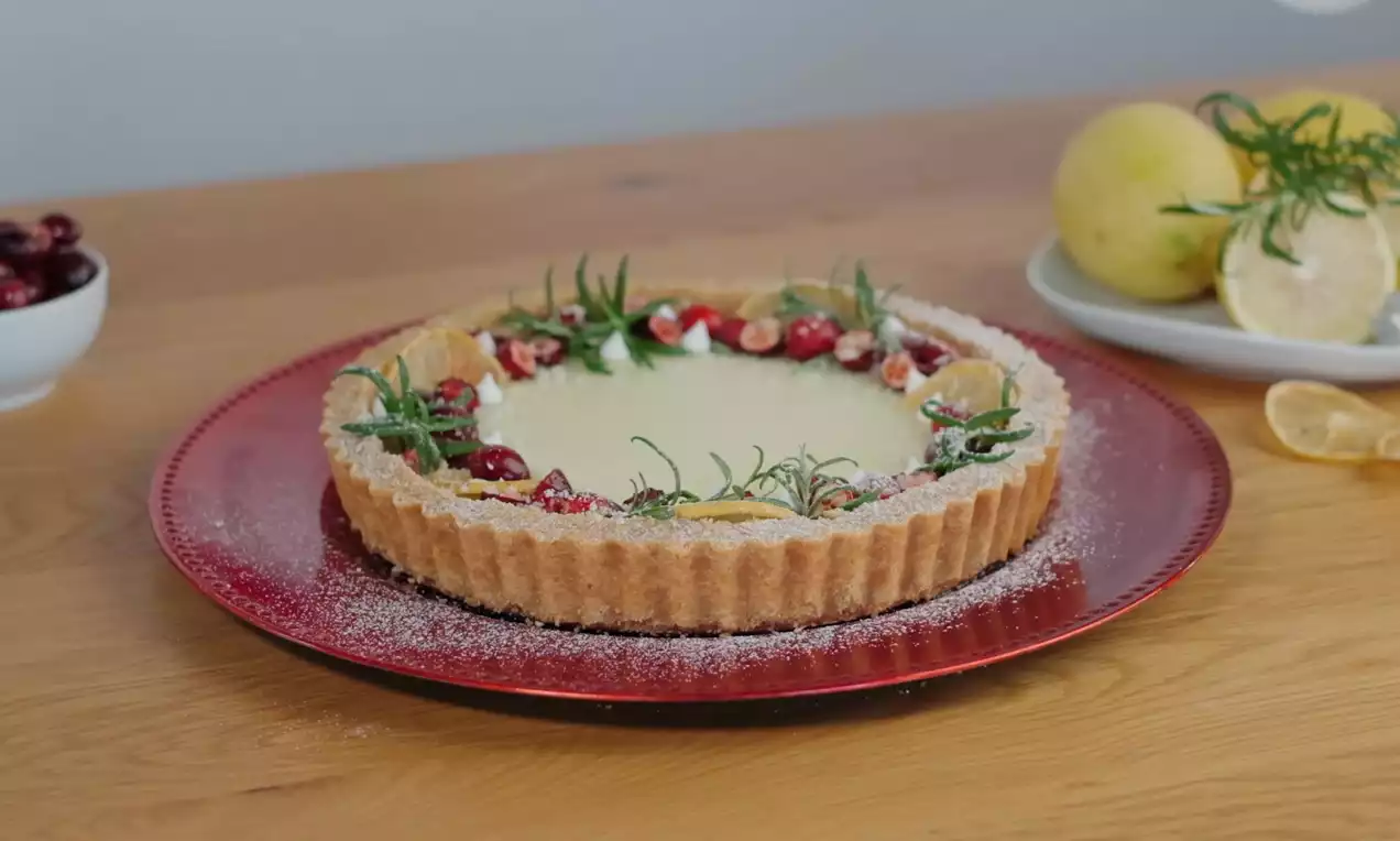 Citronový dort světlé krémové náplně v zlatavé vlnkované krustě, obložený brusinkami, cukrem a rozmarýnem na červeném talíři