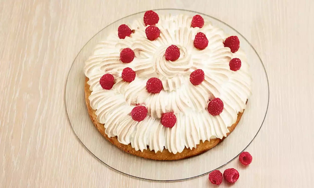 Torta alla base dorata con soffice meringa bianca a spirali guarnita da lamponi rossi su piatto trasparente e sfondo in legno chiaro