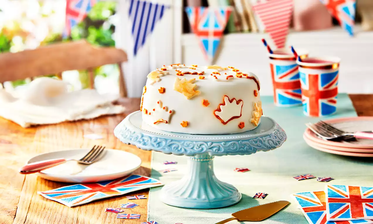 White Royal Iced Fruit Cake with smooth icing, gold and orange icing decorations, set on a light blue stand surrounded by Union Jack-themed items