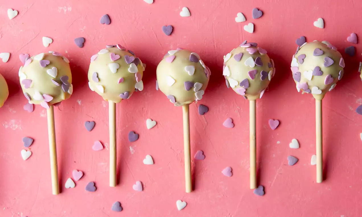 Roomkleurige cakepops op houten stokjes met een laagje glazuur en pastelkleurige suikerhartjes op een roze ondergrond