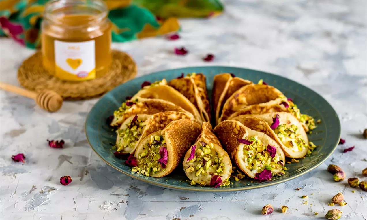 Елегантно наредени малки златисти палачинки, пълнени с гладък ванилов пудинг, украсени с натрошени шамфъстъци и розови листенца върху синя чиния