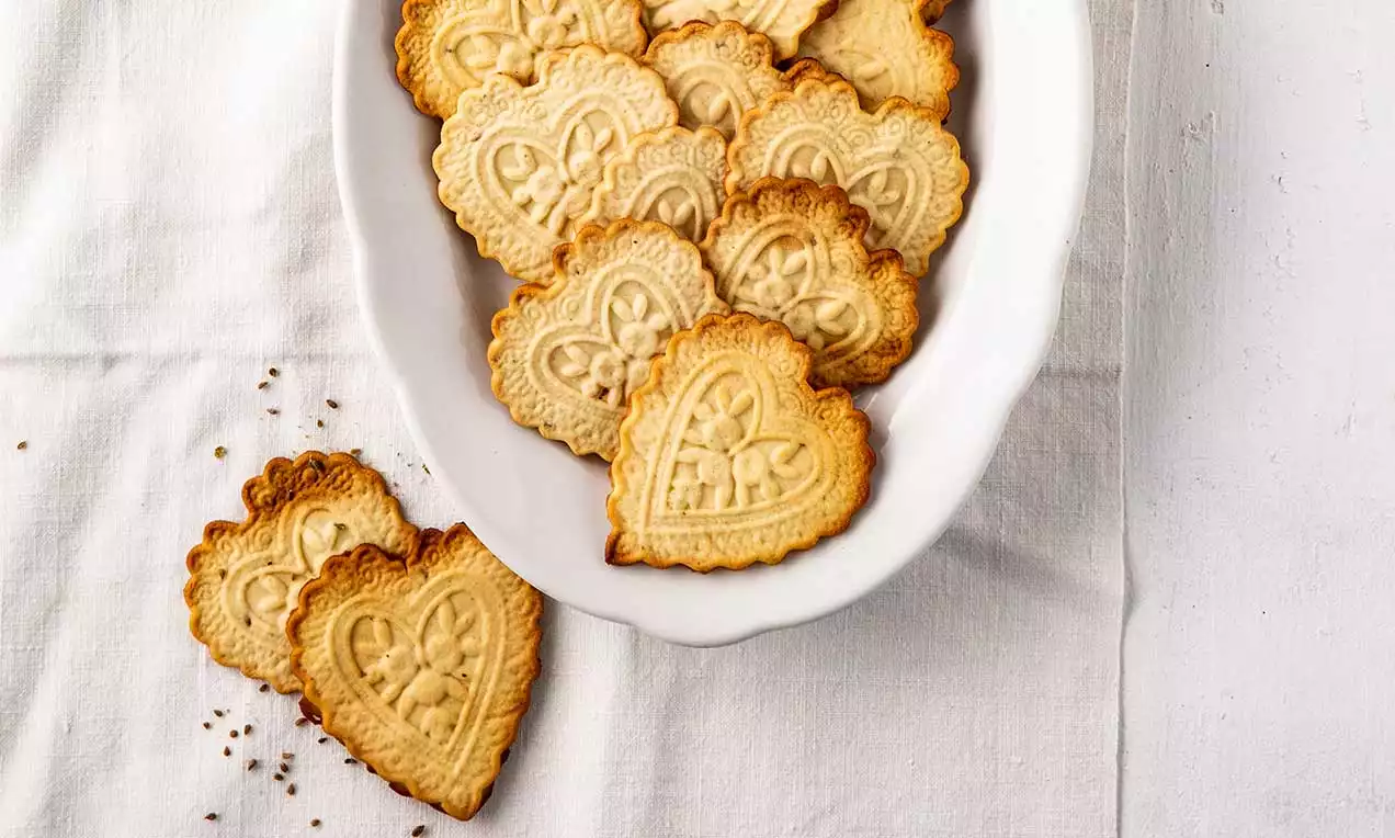 Herzförmige, goldgelbe Züri Tirggel mit kunstvollen Reliefmustern in weißer ovaler Porzellanschale auf heller Stoffunterlage