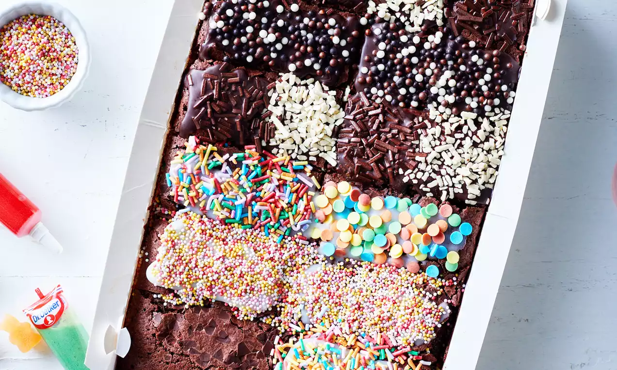 Donkerbruine brownies met glanzende chocoladetopping, felgekleurde hagelslag, witte chocoladevlokken en decoratie in een witte doos