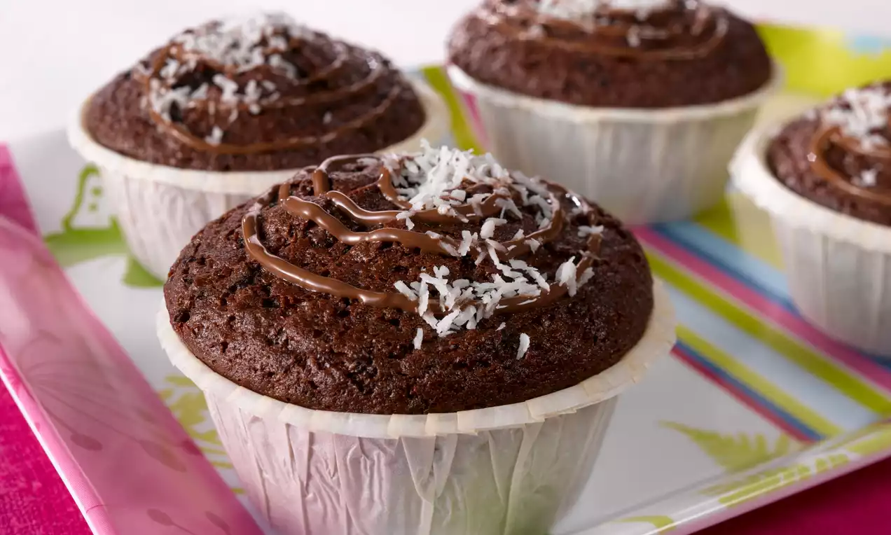 Tummanruskeita suklaamuffineja valkoisessa paperivuoassa, pinnalla kiiltävää suklaakuorrutusta ja ohuita kookoshiutaleita raidallisella tarjottimella