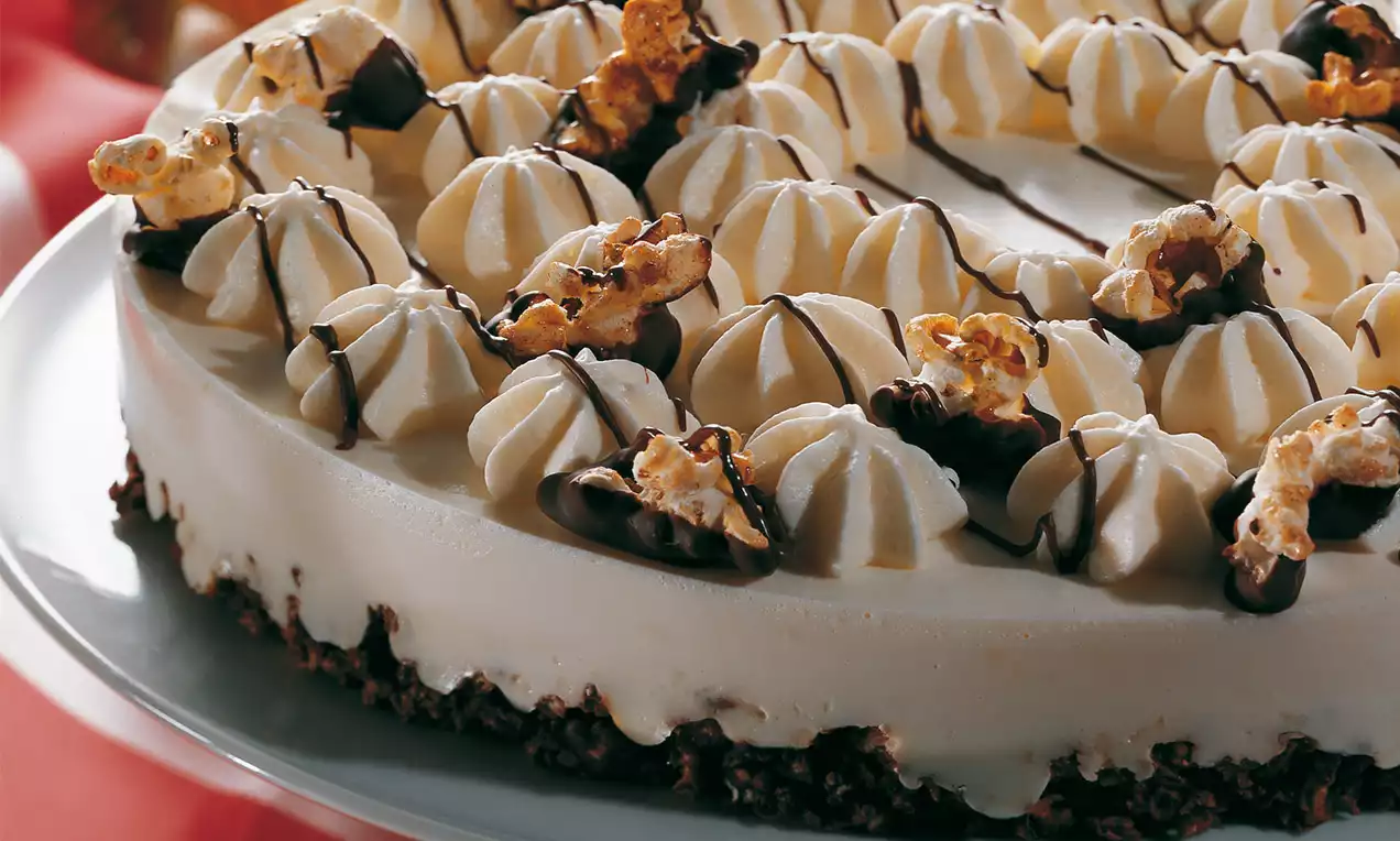 Knusprige Schokoboden-Torte mit weißer Topfencreme, Sahnetupfen, dunklen Schokostreifen und karamellisiertem Popcorn dekoriert